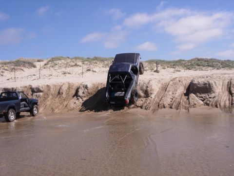 1985 Toyota Hilux - Two trucks navigate a sandy beach and climb a steep embankment under a blue sky with clouds.