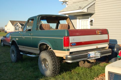1987 Ford Bronco - A lifted 1987 Ford Bronco sits on grass next to a house, with another car visible in the background.