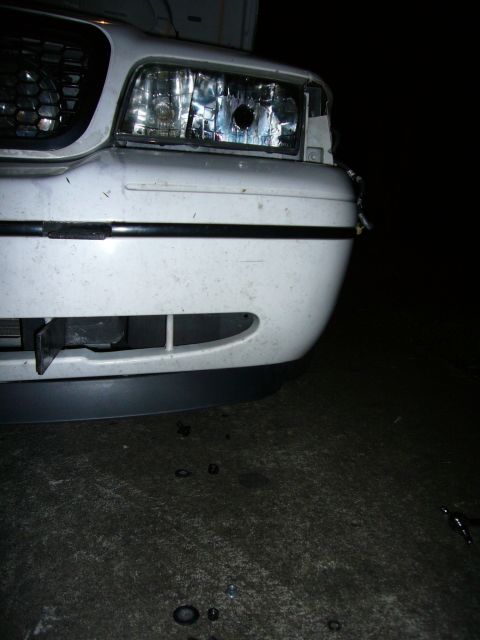 2000 Ford Crown Victoria - Night close-up of the driver's-side front corner of a white sedan, showing the composite headlight with its lens missing so the chrome refl…