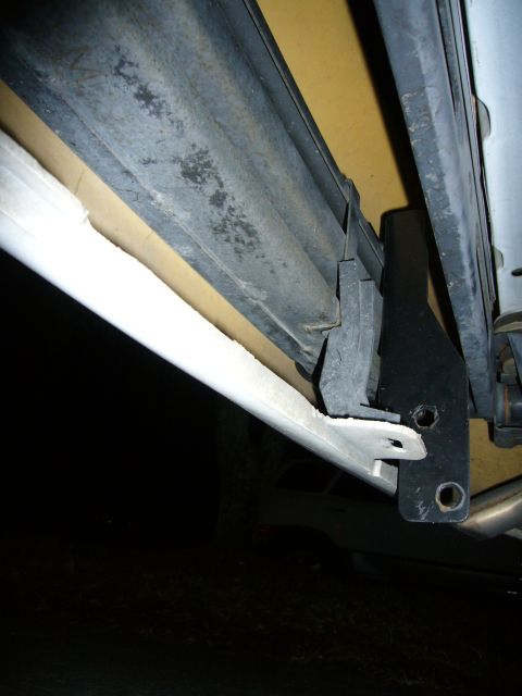2000 Ford Crown Victoria - Close-up looking up under a car's rear bumper, where a black steel bracket with two bolt holes passes through a slot cut into the lower edg…