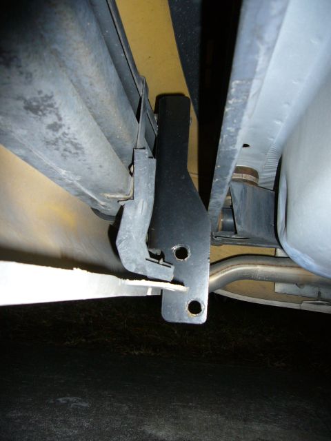 2000 Ford Crown Victoria - Low-angle underbody shot at the rear of a car, showing a black steel hitch or bracket bolted to the frame rail alongside a bare-metal mount…
