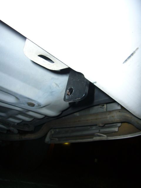 2000 Ford Crown Victoria - Flash-lit underbody view behind the rear bumper of a white car, showing a black steel bracket with a bolt hole tucked between the body pane…