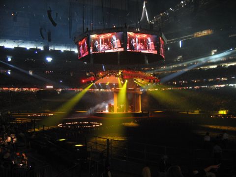 2000 Ford Crown Victoria - A live concert is underway in a stadium, with a band performing on stage and shown on multiple large screens.