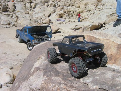 1985 Ford Bronco - RC rock crawlers navigate a challenging, rocky outdoor course with a blue pickup truck in the background.
