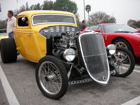1998 Ford Crown Victoria - A yellow hot rod is parked next to a red sports car, with its engine and chrome grille prominently displayed.