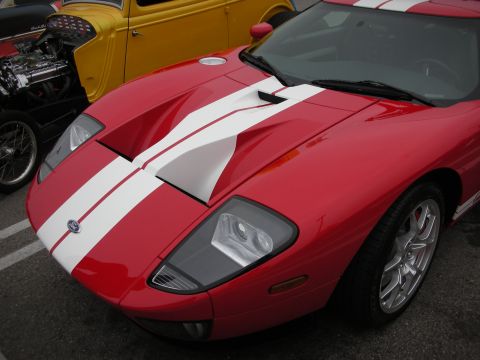 1998 Ford Crown Victoria - A red Ford GT with iconic white racing stripes is parked next to a yellow hot rod.