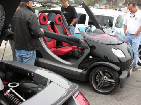1998 Ford Crown Victoria - A group of people admire a unique Smart car with its distinctive gullwing doors raised at a car meet.