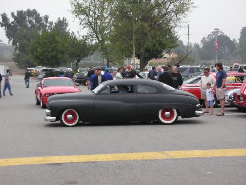 1998 Ford Crown Victoria - A matte black custom coupe with wide whitewall tires and red hubcaps is displayed at an outdoor car event.