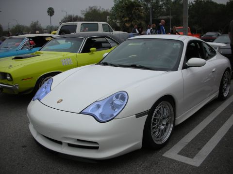 1998 Ford Crown Victoria - A white Porsche 911 is parked in a lot with other classic cars, including a yellow Plymouth Barracuda.