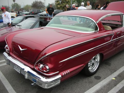 1998 Ford Crown Victoria - A shiny red 1958 classic car is parked at a meet, showing off its distinctive tail fins and chrome details.