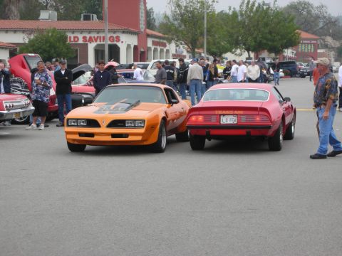 1998 Ford Crown Victoria - Two classic muscle cars, an orange one and a red one, are parked at an outdoor event with people around.