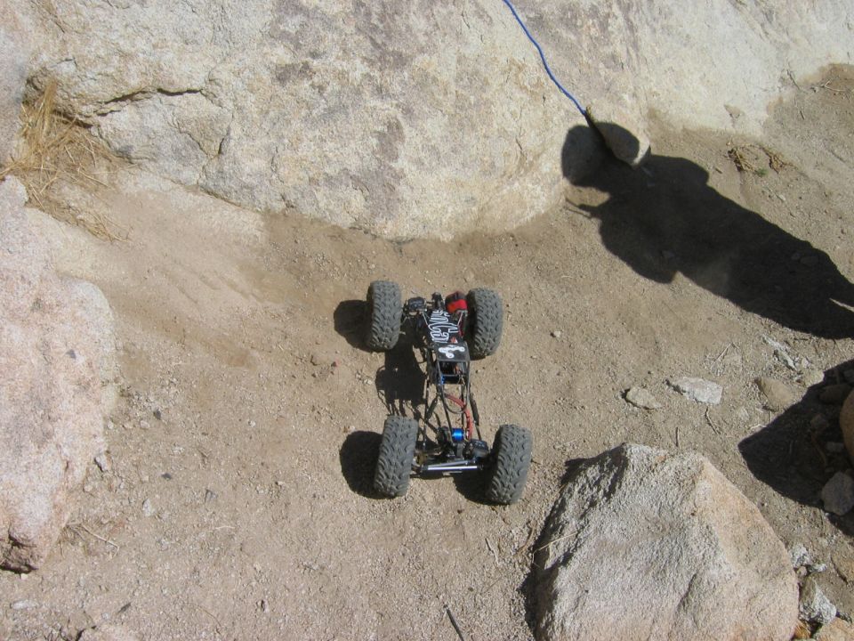 Black RC rock crawler with knobby tires on dirt among boulders, top-down view