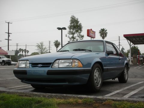 1990 Ford Mustang - This blue 1990 Ford Mustang coupe is parked in a lot, with the 'MUSTANG' text clearly visible on the front.