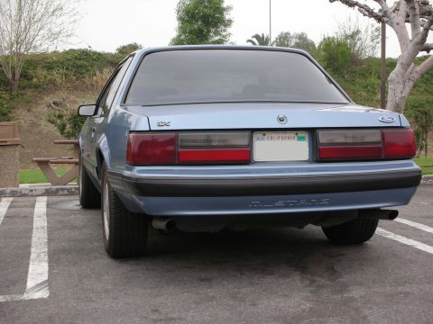 1990 Ford Mustang - The rear of a light blue 1990 Ford Mustang is shown parked in a lot with a picnic table and trees in the background.