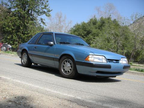 1990 Ford Mustang - A blue 1990 Ford Mustang coupe is parked on the side of a road with trees and a hill in the background.