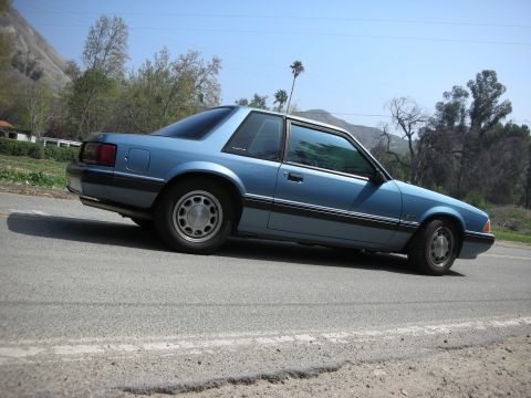 1990 Ford Mustang - Side view of a blue 1990 Ford Mustang coupe on a road with a mountainous backdrop.