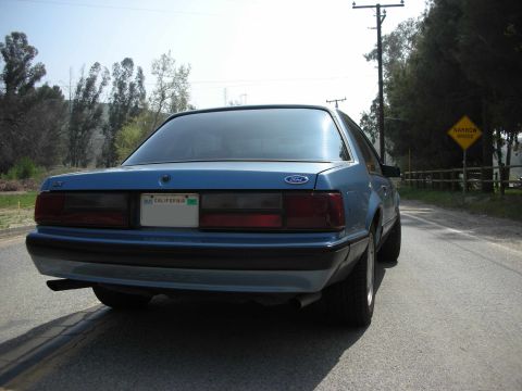 1990 Ford Mustang - A blue Ford Mustang is seen from the rear on a paved road with trees and a narrow bridge sign in the background.