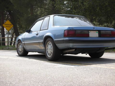 1990 Ford Mustang - Rear view of a blue coupe with a narrow bridge sign in the background.