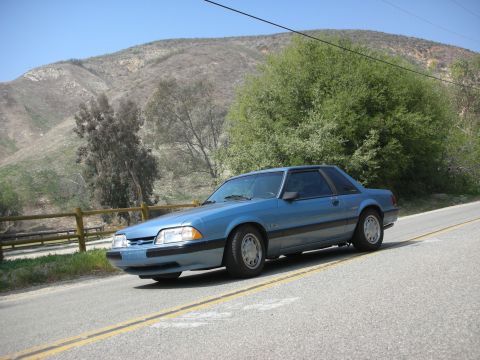 1990 Ford Mustang - A blue 1990 Ford Mustang coupe is seen driving on a road with a hillside and trees in the background.