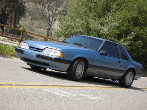 1990 Ford Mustang - A blue 1990 Ford Mustang coupe is parked on a road with a wooden fence and trees in the background.