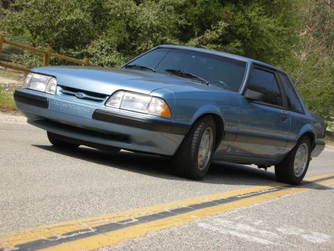 1990 Ford Mustang - This blue 1990 Ford Mustang coupe is captured from a three-quarter front view, parked alongside a road marked with double yellow lines.