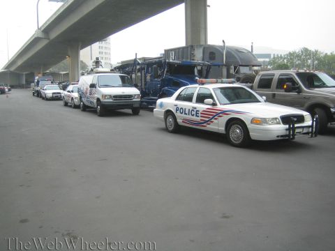 1990 Ford Bronco - White police sedan marked POLICE leads a line of emergency vehicles and trucks parked under a concrete overpass.