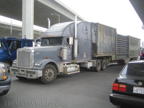 1990 Ford Bronco - Weathered gray Freightliner sleeper-cab semi truck hauling a trailer, parked beside a black VW sedan under an overpass.
