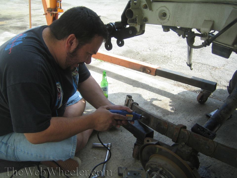 Man using rotary tool on rusty leaf spring and axle under body lifted on hoist