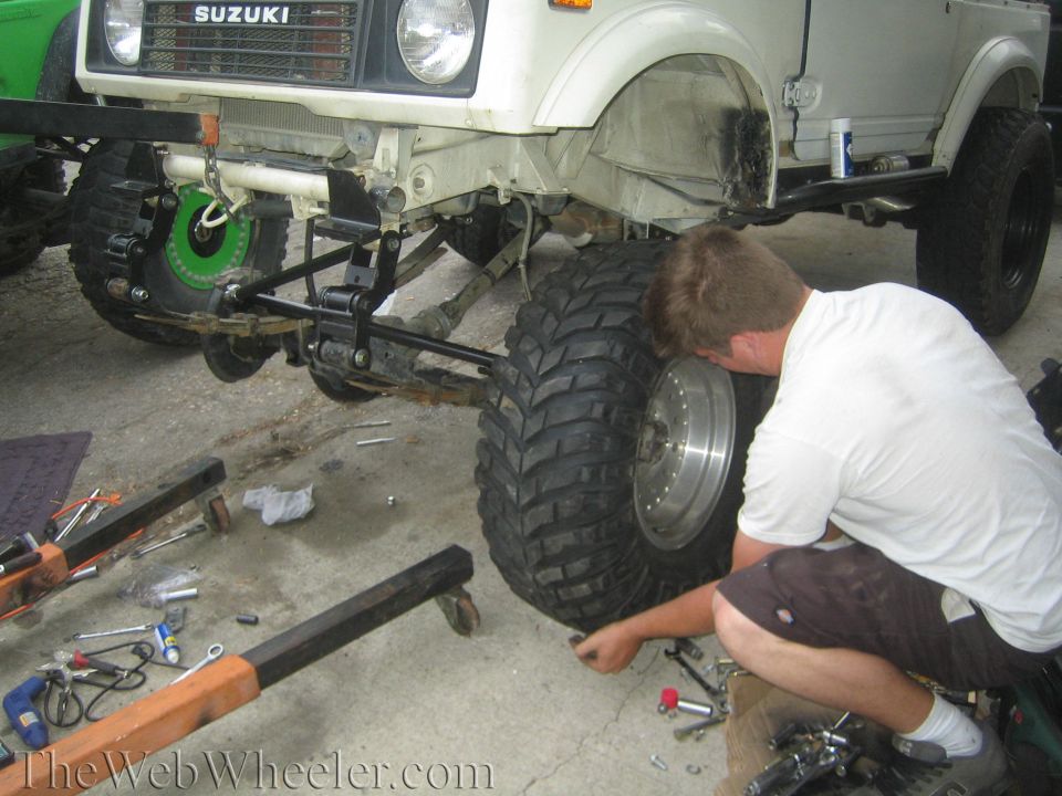 Person working on the front suspension of a lifted white Suzuki Samurai with tools scattered on concrete