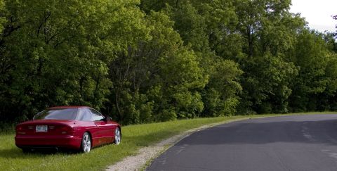 1993 Ford Probe - A red 1993 Ford Probe is parked on the grass beside a paved road, with a dense line of trees in the background.