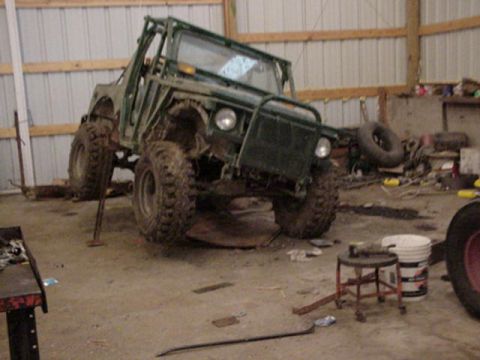 1992 Ford Bronco - A small green soft-top 4x4 with an exposed tube roll cage, no doors and mud-caked knobby tires sits flexed up on a jack stand inside a meta…