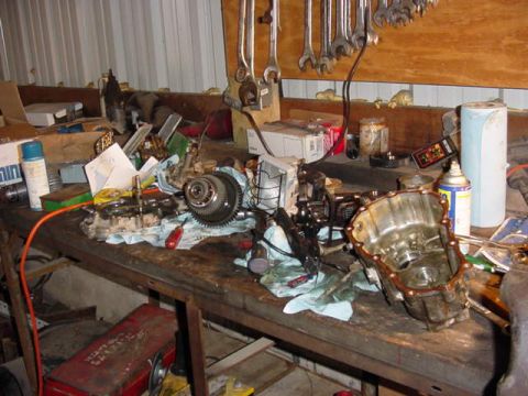 1992 Ford Bronco - A transfer case torn down across a steel workbench in a metal-sided shop, with the cast aluminum case half laid open on its side at right a…