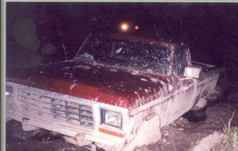 1992 Ford Bronco - Muddy red pickup truck appears stuck in the dark, possibly an early 1980s model.