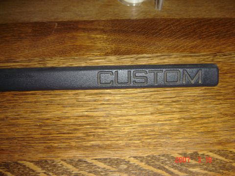 1993 Ford Bronco - A black trim piece with the word "CUSTOM" embossed on it is shown against a wooden background.