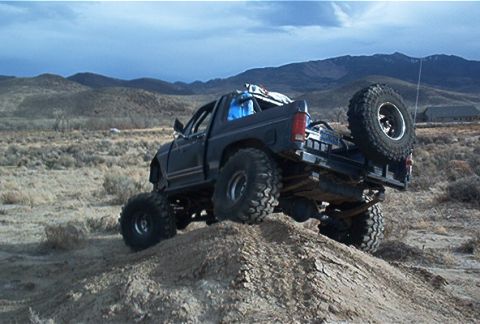 1986 Ford Bronco - This heavily modified truck, likely from the 1980s, navigates an off-road obstacle with its spare tire prominently displayed on the tailgat…