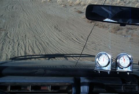 1986 Ford Bronco - The view from the driver's seat shows tire tracks in the sand and two gauges mounted on the dashboard.