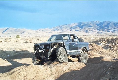 1986 Ford Bronco - A heavily modified blue truck navigates a sandy, hilly landscape under a clear sky.