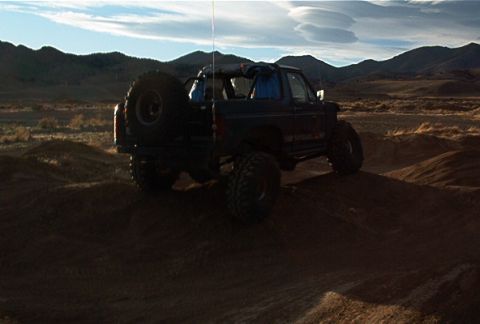 1986 Ford Bronco - A lifted Ford Bronco navigates a sandy terrain under a partly cloudy sky.