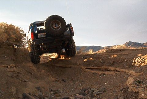 1986 Ford Bronco - A blue SUV navigates a steep dirt incline in a mountainous, arid landscape.