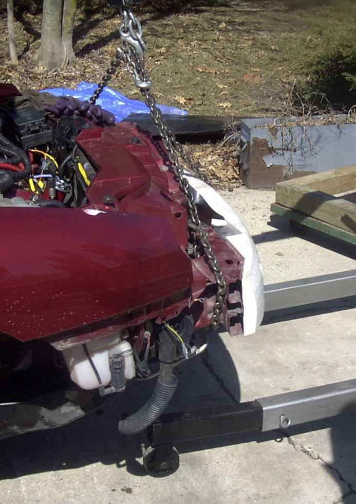 Maroon car front end suspended by chains, showing engine components and damaged headlight area