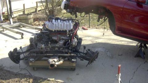 1997 Ford Taurus - A complete V6 engine and subframe assembly sits on a wooden dolly in a driveway, still carrying the rack-and-pinion steering, exhaust manif…