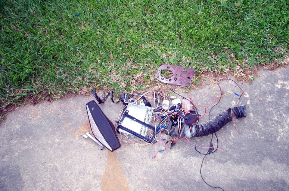 Pile of vehicle wiring, a black taillight assembly, and a rusted metal bracket on concrete next to grass