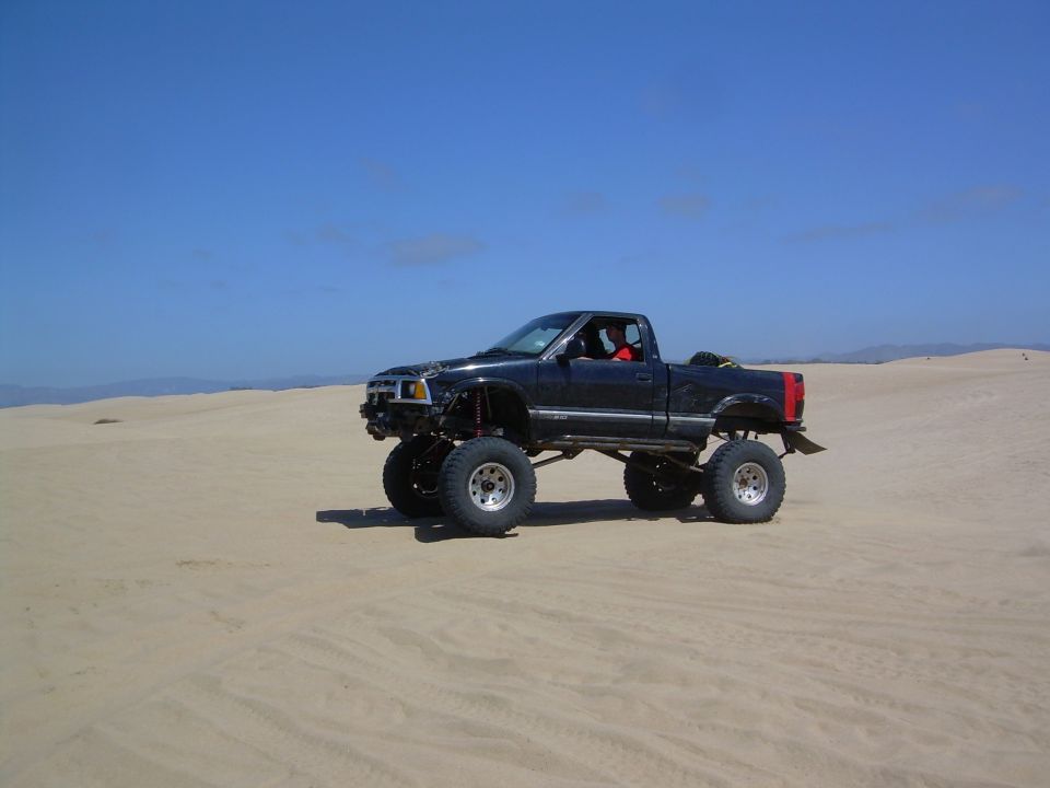 Lifted black pickup truck with large tires driving on sand dunes under a clear blue sky