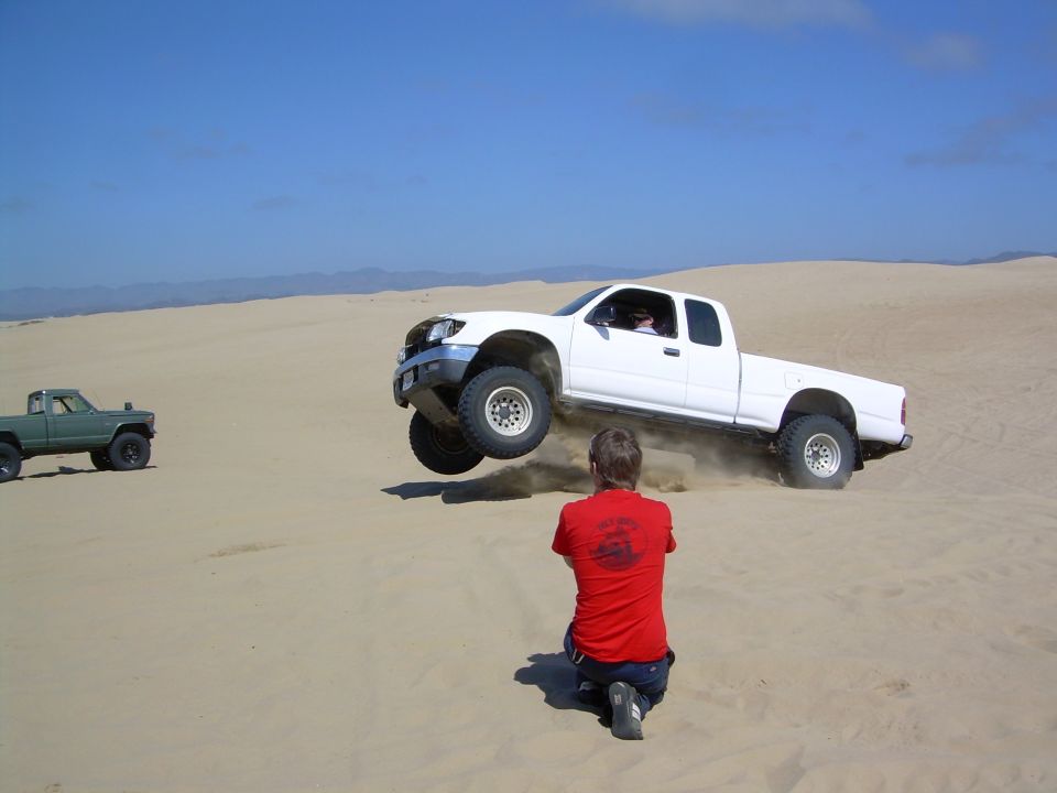 White pickup truck mid-air over sand dune, photographer kneeling. Grille suggests 2000s