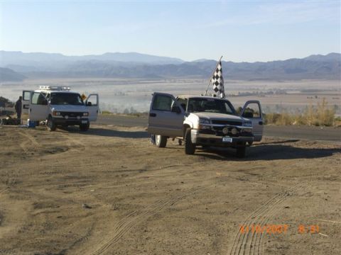 1990 Ford Bronco - Silver pickup and white van with doors open and a checkered flag, parked on a dusty desert overlook with mountains.