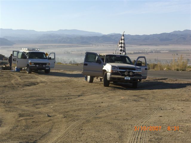 Pickup and van parked on a dirt overlook with a checkered flag and desert valley beyond