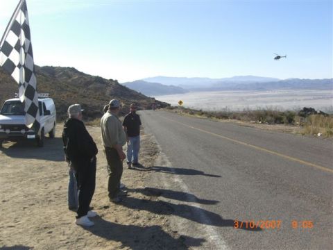 1990 Ford Bronco - Spectators stand by a checkered flag and white van along a desert road as a helicopter hovers over the hazy valley.