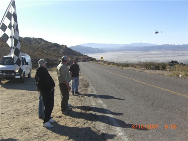 People standing roadside watching a helicopter over a desert valley