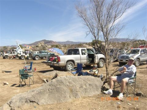 1990 Ford Bronco - Off-road desert camp with a silver pickup, checkered flags, lawn chairs and a seated man under a leafless tree.