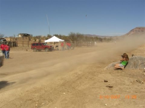 1990 Ford Bronco - Thick dust trails along a dirt course past a white tent and red SUV while a girl lies on a rock photographing the action.